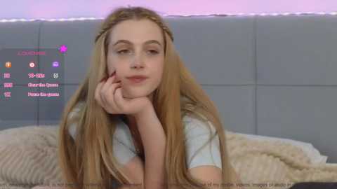 Media: Video of a young woman with long blonde hair, resting her chin on her hand, in a light grey shirt, on a bed with beige blankets and a grey headboard, with a live streaming overlay showing her viewers.