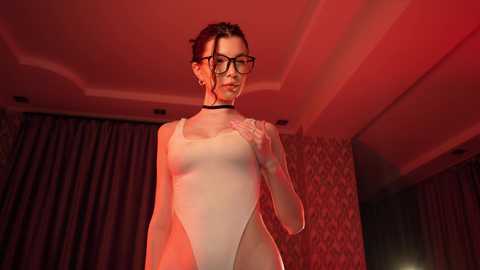 Media: Video of a slim, fair-skinned woman with dark hair in a bun, wearing black-rimmed glasses, a sheer, high-cut bodysuit, and a choker, posing in a dimly lit room with red lighting and patterned wallpaper.