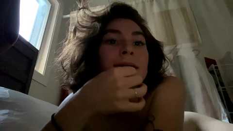 Media: Video of a young woman with light skin and shoulder-length brown hair, lying on a bed, partially covered by a white sheet. She has a neutral expression and is touching her mouth with her left hand. The background shows a window and a white curtain.