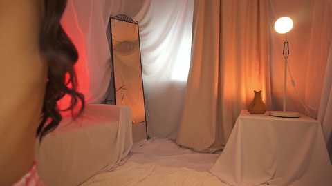 Media: Video of a dimly-lit, cozy room with beige curtains, a large mirror, a white table, a ceramic cat, and a soft lamp casting warm light, creating a serene, intimate atmosphere.