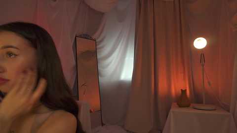 Media: A dimly lit video featuring a woman with long dark hair, wearing a white top, touching her face in a reflective mirror. A warm, ambient light illuminates a small table with a vase. The background has a sheer curtain.