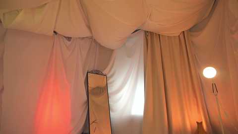 Media: Video of a dimly lit room with white drapes, a mirror, and a round lamp casting a warm glow, creating a cozy, intimate atmosphere.