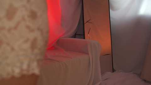 Media: Video of a beige armchair partially covered with white fabric, illuminated by a red light, set in a dimly lit room with a mirrored wall and a warm orange glow from a nearby lamp.