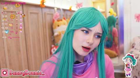 Media: A colorful video of a woman with long turquoise hair and a pink top, wearing a distressed expression, surrounded by plush toys in a bright room.