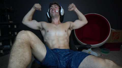 Media: Video of a muscular, shirtless man with headphones, flexing his biceps while sitting on a carpeted floor, wearing blue shorts, in a dimly lit room with a red chair and shelves in the background.