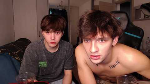 Media: Video of two young men with pale skin, one shirtless, sitting at a gaming desk in a dimly lit room. They appear to be playing video games.