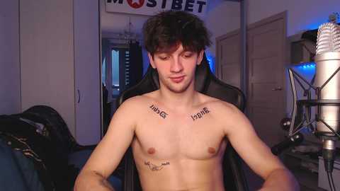 Media: Video of a shirtless, light-skinned, young man with short, dark hair, sitting on a black gaming chair, wearing tattoos with \"HOME\" on his chest and \"HUGE\" on his shoulder, in a dimly lit room with gaming equipment.