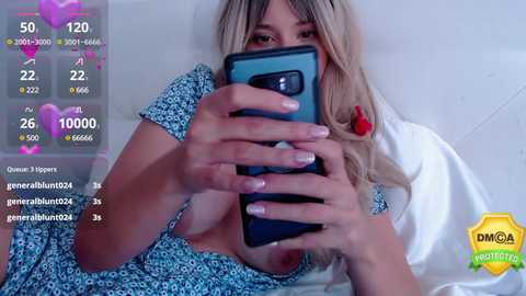 Media: Video of a blonde woman with long hair, wearing a blue floral dress, taking a selfie in a mirror. She holds a smartphone, showing a heart emoji on the screen.