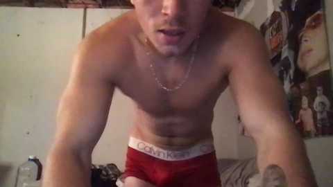 Media: Video of a shirtless, fair-skinned young man with a slender build wearing red Calvin Klein briefs, leaning forward in a dimly lit room. Background includes posters and a messy room.