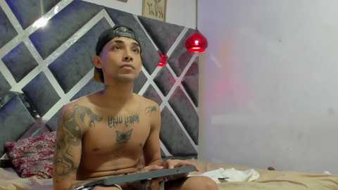 Media: Video of a shirtless, tattooed young man in a bed, wearing a backward cap, with a laptop, in a dimly lit, modern bedroom with geometric-patterned headboard and red pendant lights.