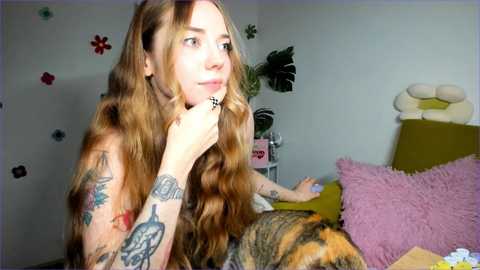 Media: Video of a young woman with long, wavy brown hair, tattoos on her arms, and a contemplative expression, sitting on a bed in a brightly lit room with floral wall stickers and plush toys.