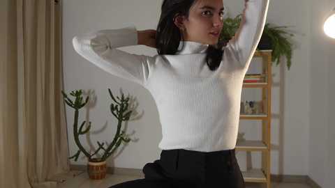 Media: Video of a young woman with medium-length black hair, wearing a white ribbed turtleneck and black pants, stretching in a modern, minimalistic living room with beige curtains, potted plants, and wooden shelves.