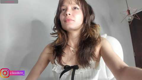 Media: A video of a young Asian woman with shoulder-length brown hair, wearing a white, ruffled, strapless top. She has a neutral expression, and the background is a plain, light-colored wall.