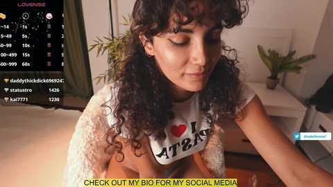 Media: Video of a curly-haired woman in a white \"I \u2764\ufe0f ATR\" shirt, standing in a living room with a TV screen displaying social media statistics.