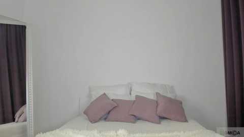 Media: A minimalist bedroom video featuring a white bed with three pillows, two in white and one in pink, against a plain white wall. Dark purple curtains frame the scene on both sides.