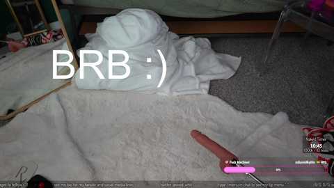 Media: Video of a bedroom with a white towel on the floor, a mirror reflecting part of the bed, a wooden dresser, and a red dildo. Text reads \"BRB.\