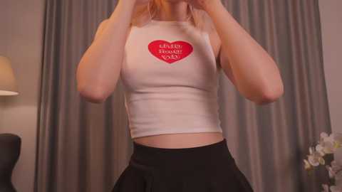 Media: Video of a slim, light-skinned woman with blonde hair, wearing a white crop top with a red heart and \"HAPPY BIRTHDAY\" text, and black high-waisted skirt, against gray curtains in a modern room.