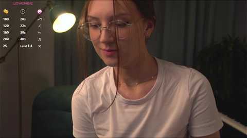 Media: Video of a young woman with fair skin, brown hair, and glasses, wearing a white T-shirt, in a dimly lit room with a green chair and hanging light bulb.