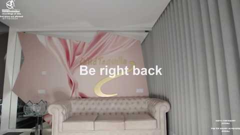 Media: Video of a living room with a beige tufted sofa, pink floral wall mural, and white vertical blinds. Text overlay reads, \"Be right back.\