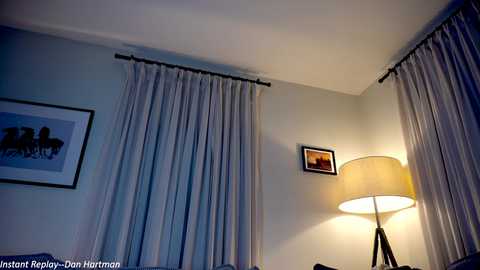 Media: Video of a dimly lit bedroom with beige walls, a tall floor lamp, and sheer grey curtains. A framed photo of a horse and rider hangs on the left wall.