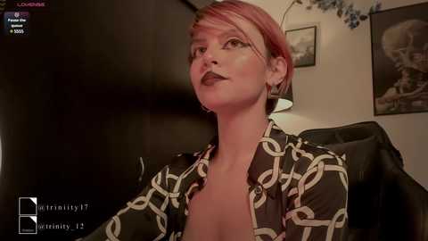 Media: Video of a woman with short, pink hair, dark lipstick, and a black-and-white patterned blouse, seated in a dimly lit room with a skull-themed painting on the wall.