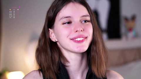 Media: Video of a young Caucasian woman with fair skin, straight brown hair, and a slight smile, wearing a black top, in a softly lit room with blurred background.