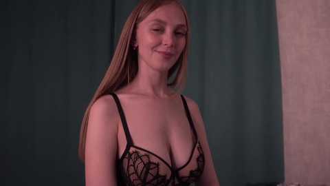 Media: Video of a smiling, fair-skinned, blonde woman with medium-sized breasts wearing a sheer black lace bra. She stands against a dark teal curtain and a beige wall, creating a subtle contrast.