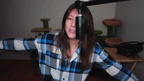 Media: Video of a woman with long brown hair, wearing a blue plaid shirt, playing a black guitar with a green guitar stand in the background.