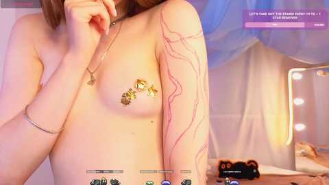 Media: A topless woman with fair skin and brown hair, wearing a gold necklace with a flower pendant, poses in a soft, pink-lit room. She has a tattoo of a snake on her right arm.