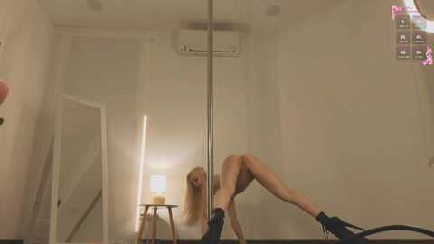 Media: Video of a nude, slender, blonde woman with long hair performing a pole dance in a dimly lit, minimalist room with white walls, a mirror, and a white door.