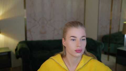 Media: Video of a blonde woman with light skin, wearing a yellow hoodie, posing indoors. She has long hair tied back, and is adorned with large, dangling earrings. Background features a green velvet couch, beige walls with a floral pattern, and a lamp on a side table.