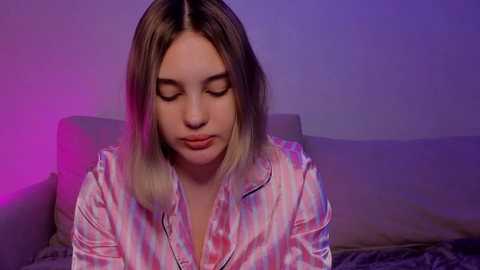 Media: A video of a young woman with straight, shoulder-length blonde hair, wearing a pink and white striped satin shirt. She has a calm, contemplative expression, sitting on a bed with a soft, blurred background.