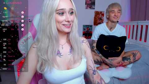 Media: Video of a smiling, blonde, tattooed woman in a white tank top, sitting on a bed with a man holding a black cat. Background includes pink and white decor, a gaming chair, and a TV.