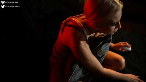 Media: Video of a blonde woman in a red dress, crouched, holding a tablet, in a dimly lit room, with a watermark in the corner.