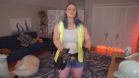 Media: Video of a plus-sized woman with blue hair and a white tank top, wearing a neon yellow vest and blue shorts, holding a camera, standing in a modern living room with a plush rug, a blue couch, and \"Astro Ocean\" neon sign.