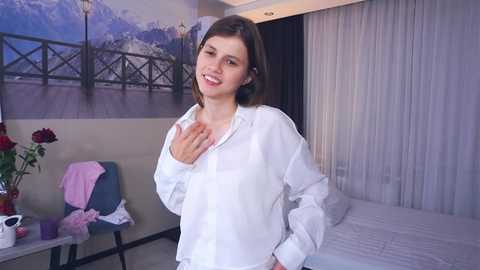 Media: Video of a smiling young woman with shoulder-length brown hair in a white shirt, standing in a modern bedroom with a white bed, floral wall art, and a blue chair.