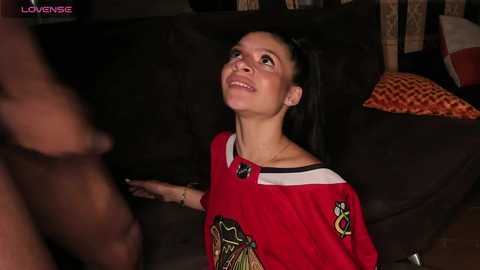 Media: Video of a smiling, light-skinned woman with long dark hair in a red off-shoulder shirt, seated on a dark sofa, dimly lit living room background, blurred man standing.