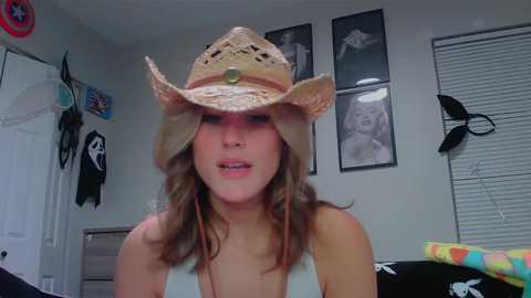 Media: Video of a young woman with light skin and wavy brown hair, wearing a beige cowboy hat and a sleeveless white top, standing indoors amidst a cluttered room with posters, a backpack, and a Thor helmet.