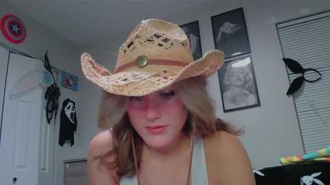 Media: Video of a blonde woman wearing a straw cowboy hat, with a white tank top, in a cluttered bedroom with posters, a door, and a window.