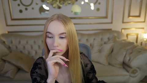 Media: Video of a blonde woman with long hair, light skin, wearing a black lace dress, thoughtfully touching her lips. Background features a plush beige sofa and decorative mirror.