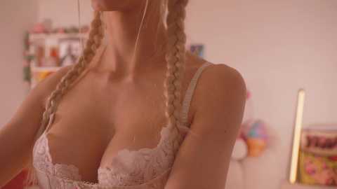 Media: Video of a blonde woman with twin braids, wearing a lacy white bra, standing indoors. Background features shelves with colorful items and a ladder. Warm lighting.