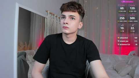 Media: Video of a young, light-skinned man with short brown hair, wearing a black t-shirt, sitting in a living room with a gray couch and sheer curtains. Virtual gaming stats overlay the image.