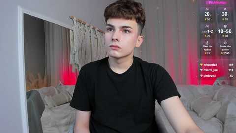 Media: Video of a young man with short, curly brown hair, wearing a black t-shirt, sitting in a living room with beige furniture and a colorful digital screen displaying social media statistics.