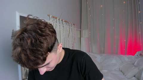 Media: Video of a young man with short, curly brown hair, shaved on one side, wearing a black t-shirt, standing in a bedroom with a white bed and beige curtains with red lighting.