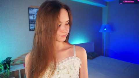 Media: Video of a young woman with long, straight brown hair, wearing a white lace-trimmed spaghetti strap top, standing in a dimly lit room with blue ambient lighting.