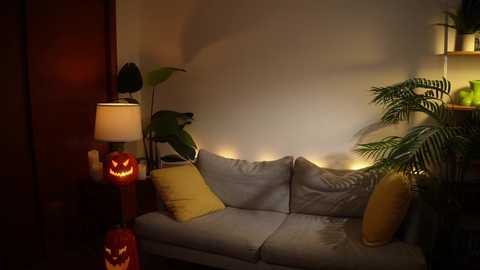 Media: Video of a cozy, dimly lit living room with a gray sofa, a lit pumpkin, potted plants, and a lamp, creating a warm, Halloween-themed atmosphere.