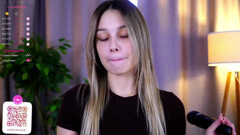 Media: Video of a young woman with long, straight, blonde hair, wearing a black top, smiling while holding a microphone, against a softly lit background with a lamp and green plant.