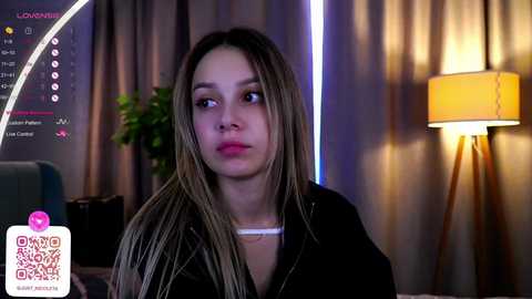 Media: Video of a young woman with long, straight, light brown hair, wearing a black top, sitting indoors, in front of a green plant and a yellow lamp.