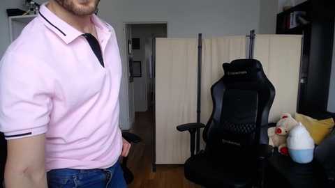 Media: Video of a man in a light pink polo shirt and jeans standing near a black gaming chair in a modern living room with beige curtains and wooden floor.