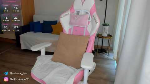 Media: Video of a pink and white gaming chair with a brown pillow, set in a modern, bright living room with white walls, a sofa, and a plant on a side table.